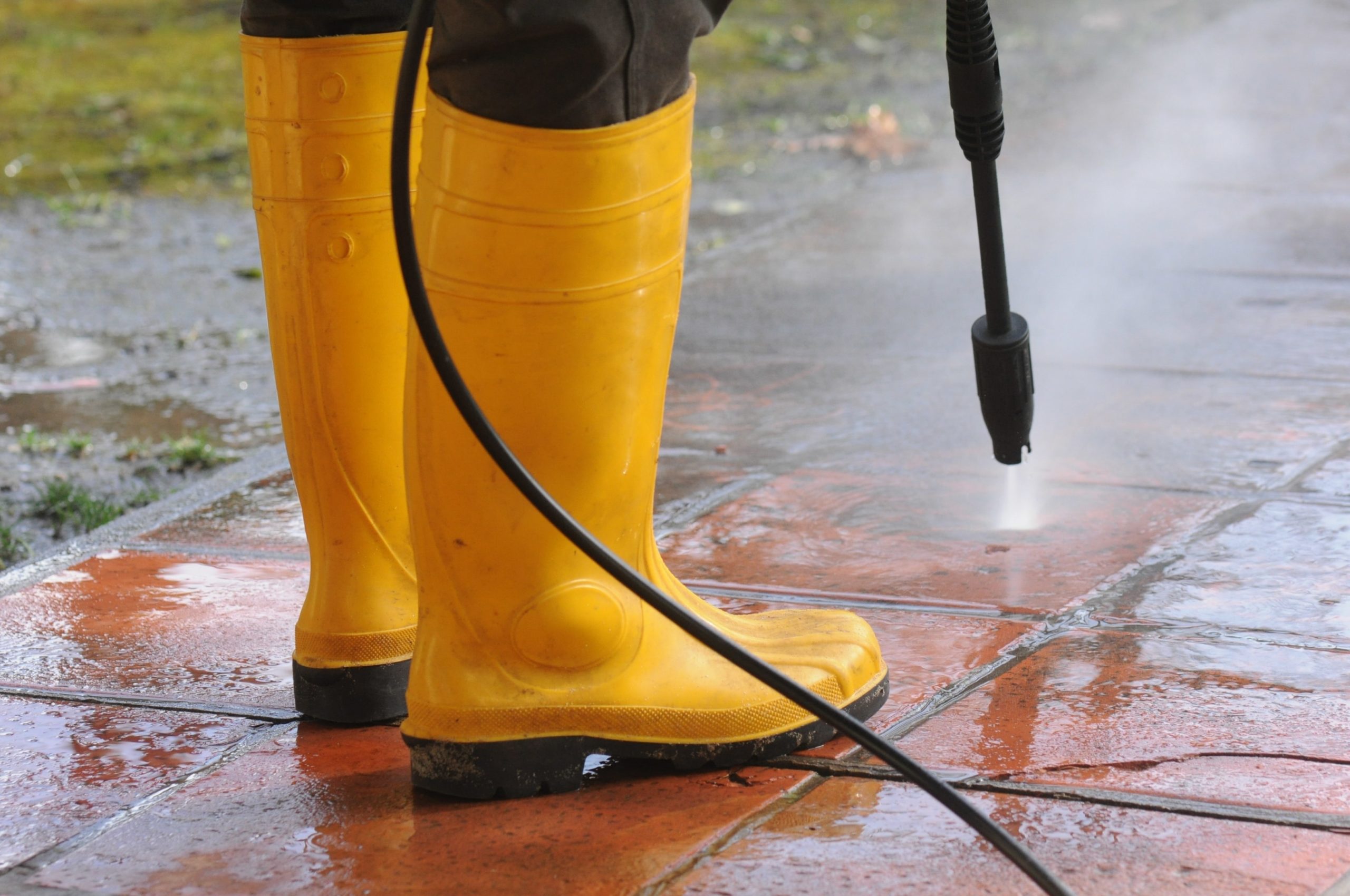person-wearing-yellow-rubber-boots-with-high-pressure-water-nozzle-cleaning-dirt-tiles-min